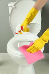 Photo of Woman with spray and rag cleaning toilet seat in bathroom, closeup