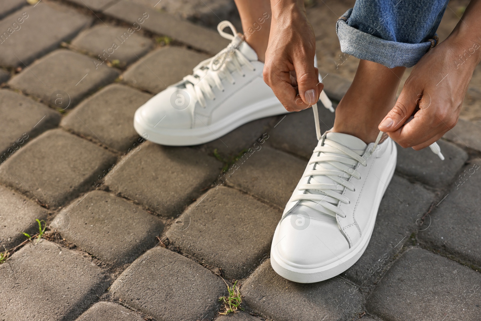 Photo of Woman tying shoelace of white sneaker outdoors, closeup