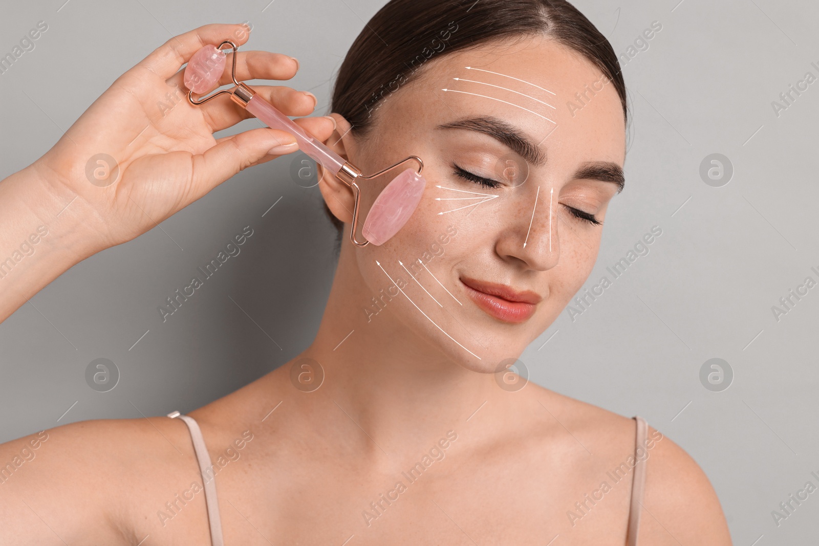 Image of Beautiful young woman doing facial massage with roller on grey background. Lines on skin showing directions of motion