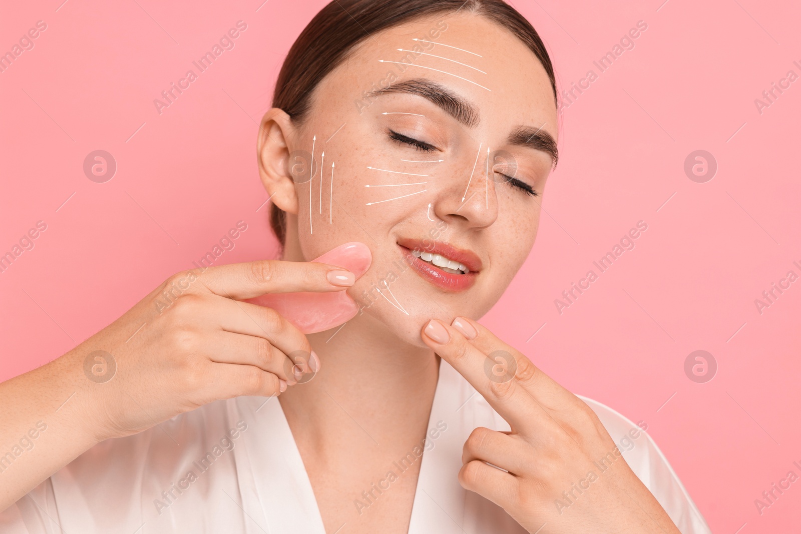 Image of Beautiful young woman doing facial massage with gua sha tool on pink background. Lines on skin showing directions of motion