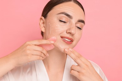 Image of Beautiful young woman doing facial massage with gua sha tool on pink background. Lines on skin showing directions of motion