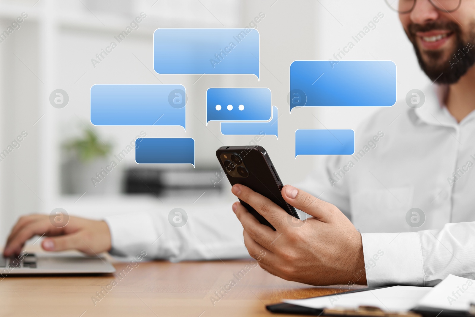 Image of Happy man reading message on smartphone at table, closeup. Speech bubbles near device