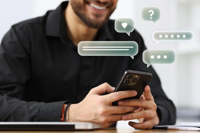 Happy businessman sending message via smartphone at table, closeup. Speech bubbles near device.