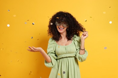 Happy young woman wearing carnival mask and confetti on yellow background