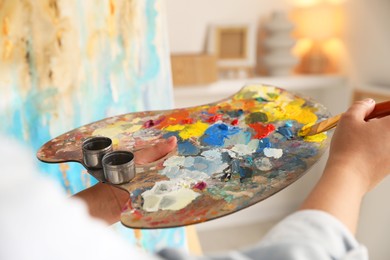 Photo of Woman mixing paints on palette with brush near easel indoors, closeup