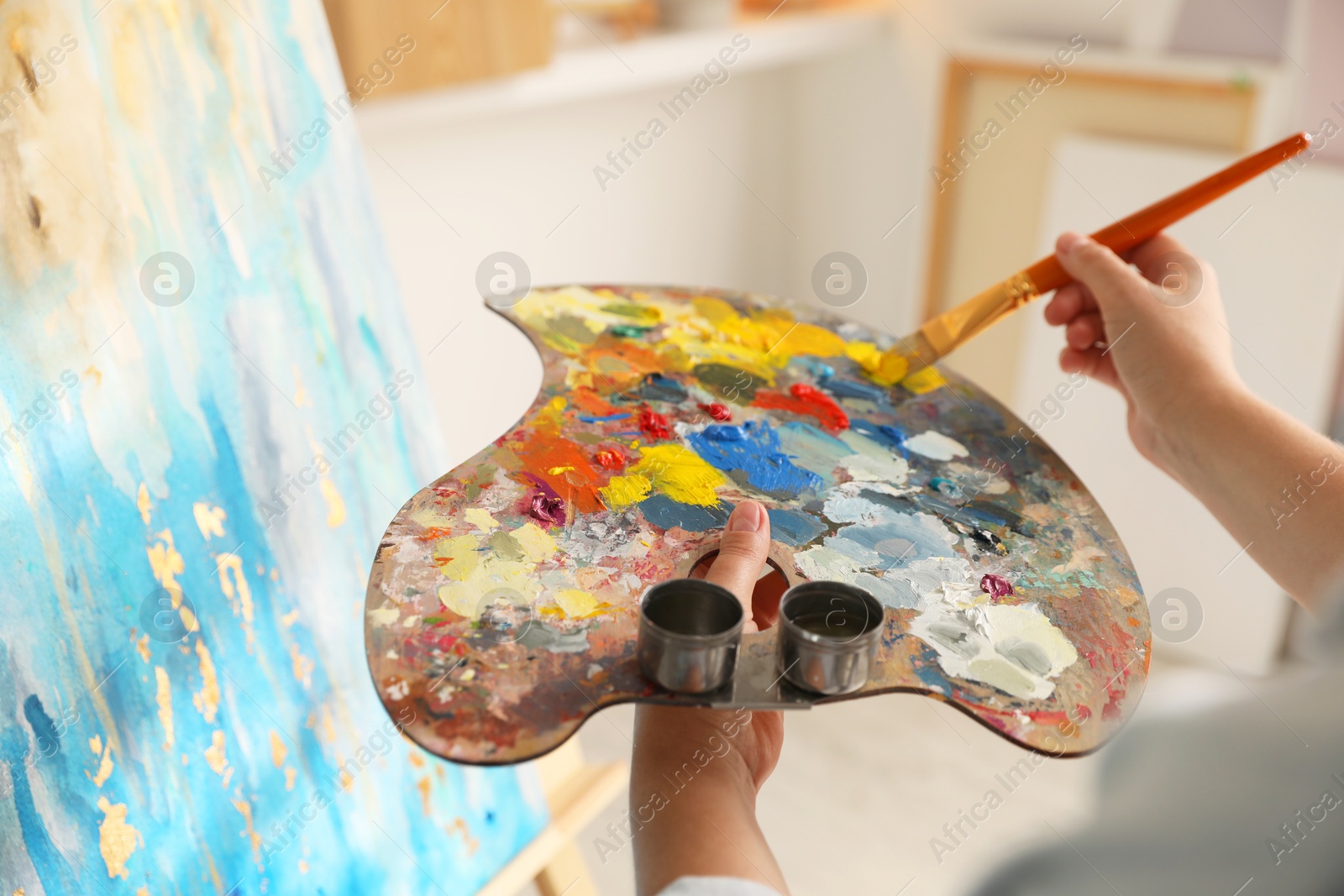 Photo of Woman mixing paints on palette with brush near easel indoors, closeup