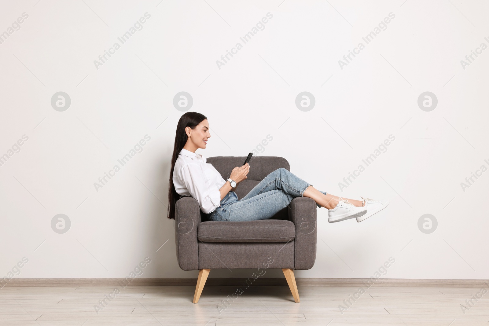 Photo of Smiling woman with smartphone sitting on armchair against white wall