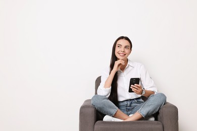 Smiling woman with smartphone sitting on armchair against white background. Space for text