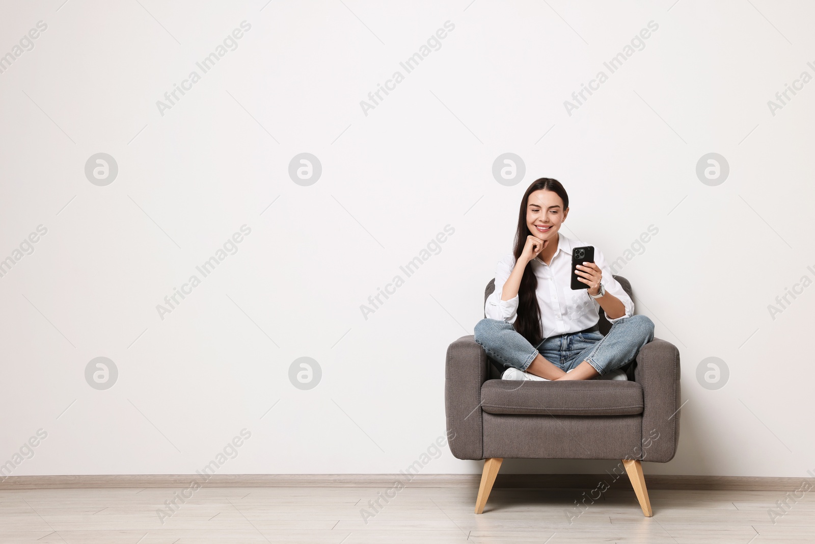 Photo of Smiling woman with smartphone sitting on armchair against white wall. Space for text