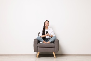 Photo of Smiling woman with smartphone sitting on armchair against white wall