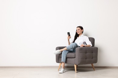 Photo of Smiling woman with smartphone sitting on armchair against white wall. Space for text