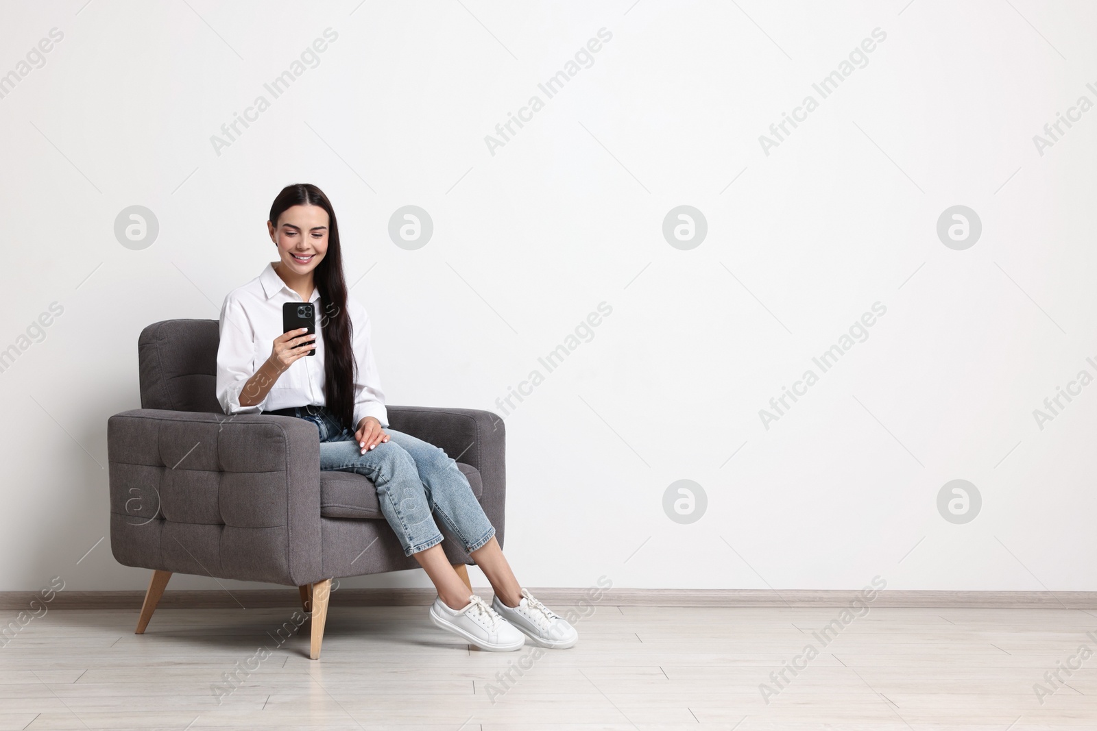 Photo of Smiling woman with smartphone sitting on armchair against white wall. Space for text