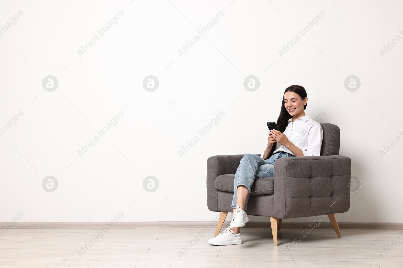 Photo of Smiling woman with smartphone sitting on armchair against white wall. Space for text