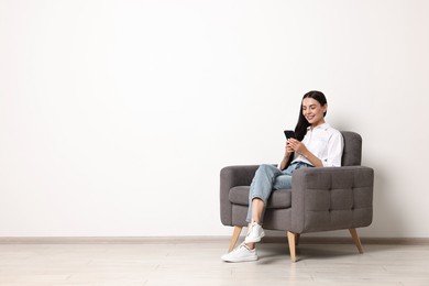 Photo of Smiling woman with smartphone sitting on armchair against white wall. Space for text