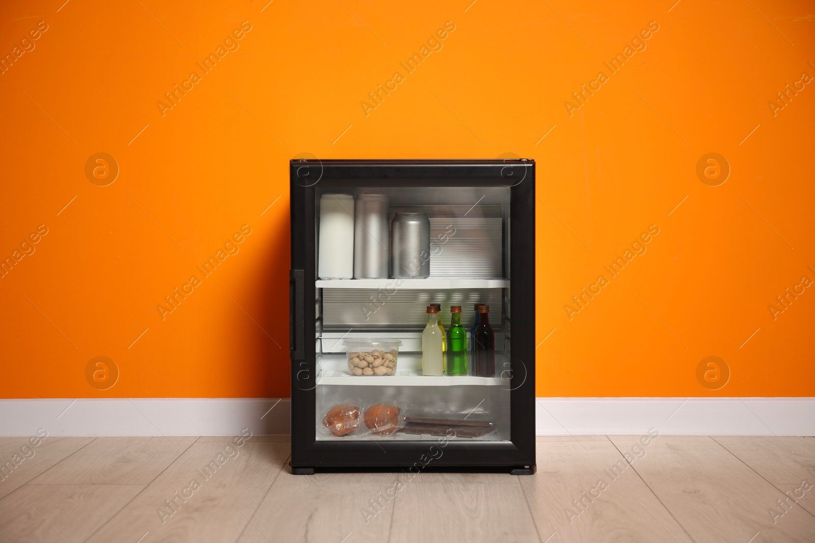 Photo of Mini refrigerator with different drinks and snacks near orange wall