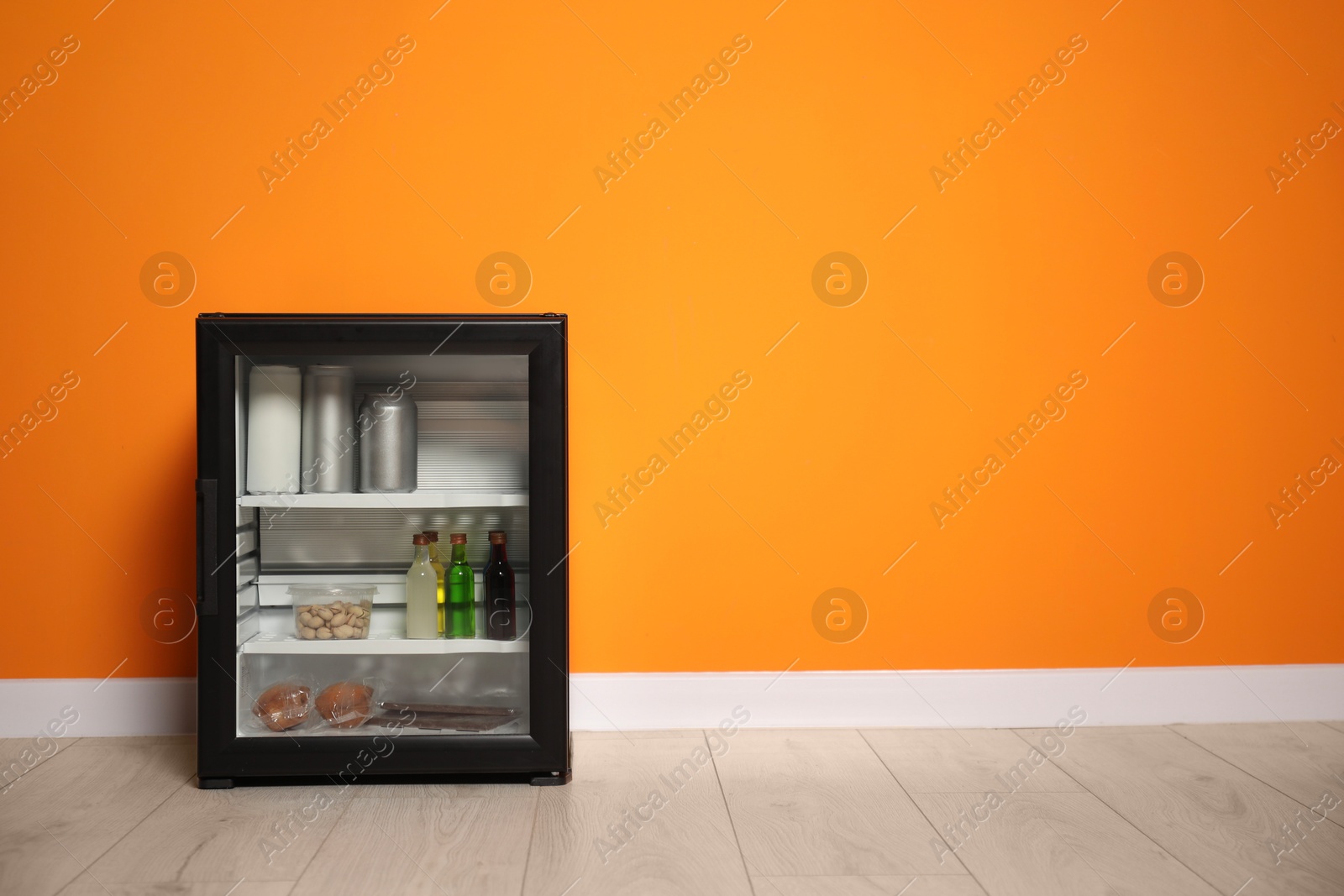 Photo of Mini refrigerator with different drinks and snacks near orange wall, space for text