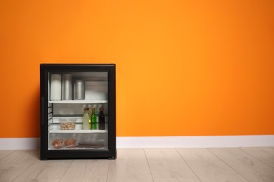 Mini refrigerator with different drinks and snacks near orange wall, space for text