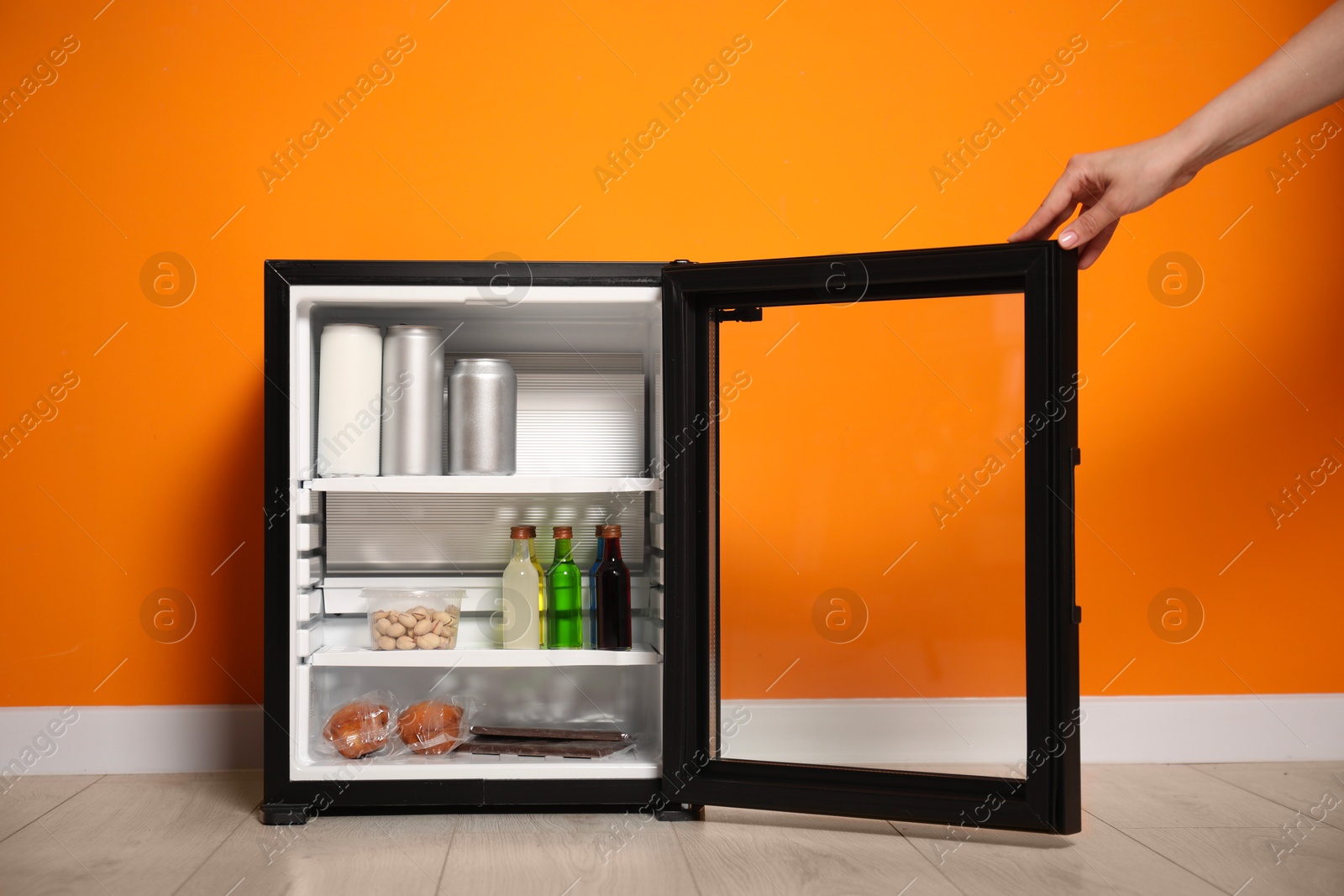 Photo of Woman opening mini refrigerator with different drinks and snacks near orange wall, closeup