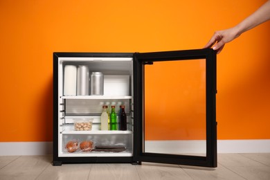 Woman opening mini refrigerator with different drinks and snacks near orange wall, closeup