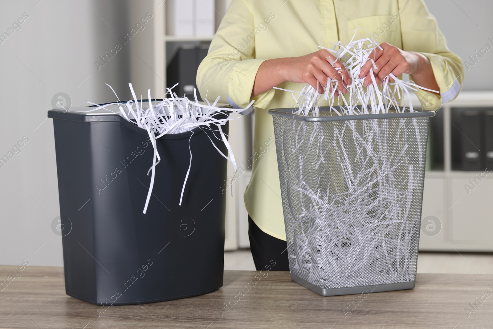 Photo of Woman putting shredded paper strips into trash bin at wooden table in office, closeup