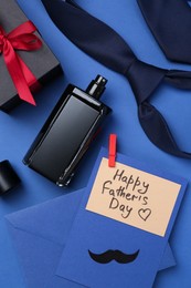 Photo of Greeting card with phrase Happy Father's Day, envelope, bottle of perfume, tie and gift on blue background, flat lay