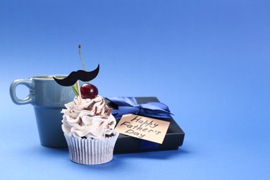 Photo of Happy Father's Day. Cupcake with mustache topper, gift box and cup on blue background. Space for text