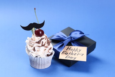 Photo of Gift box, card with phrase Happy Father's Day and cupcake with mustache topper on blue background, closeup