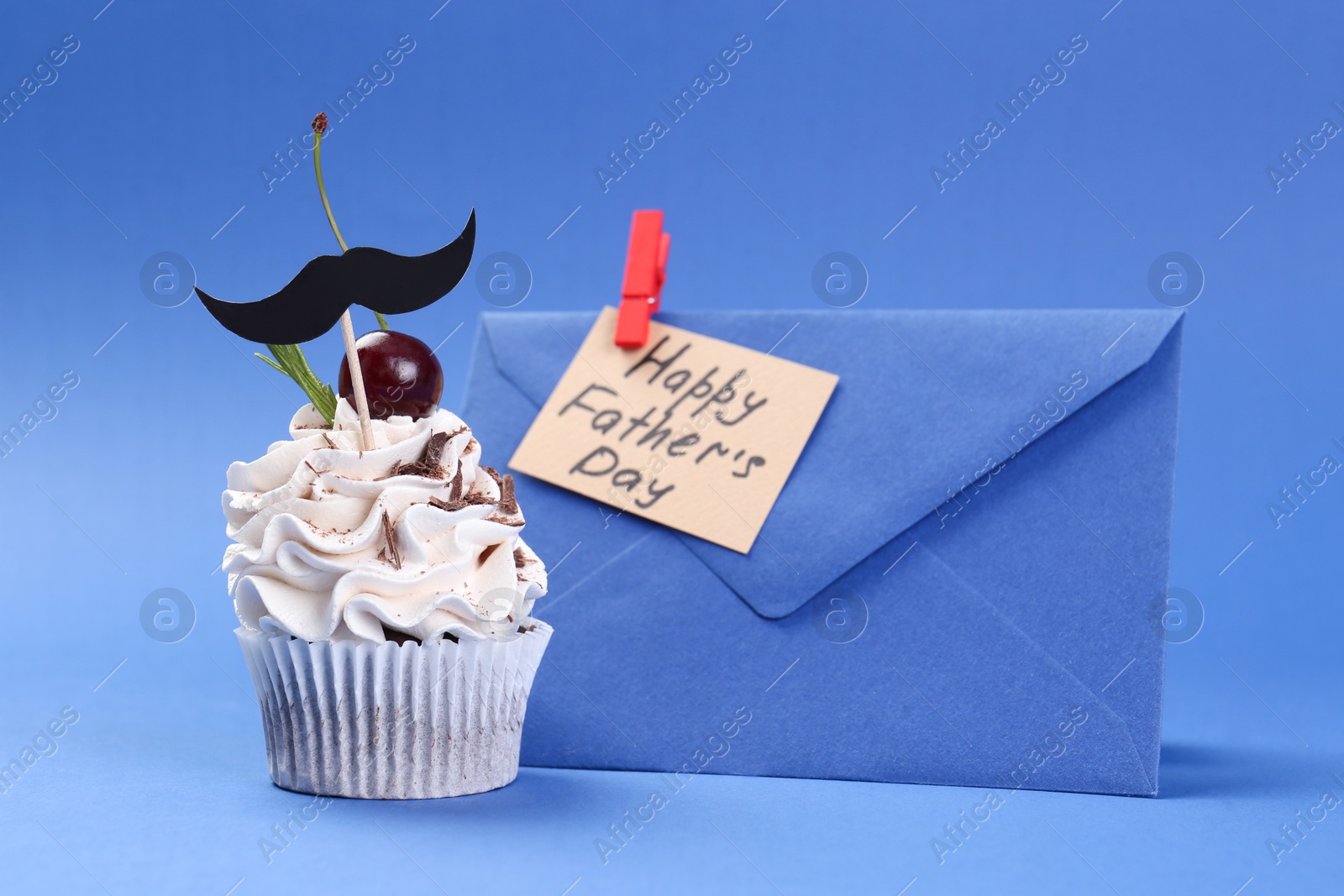 Photo of Cupcake with mustache topper, envelope and card with phrase Happy Father's Day on blue background, closeup