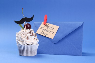 Photo of Cupcake with mustache topper, envelope and card with phrase Happy Father's Day on blue background, closeup