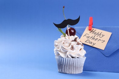 Photo of Cupcake with mustache topper, envelope and card with phrase Happy Father's Day on blue background, closeup. Space for text