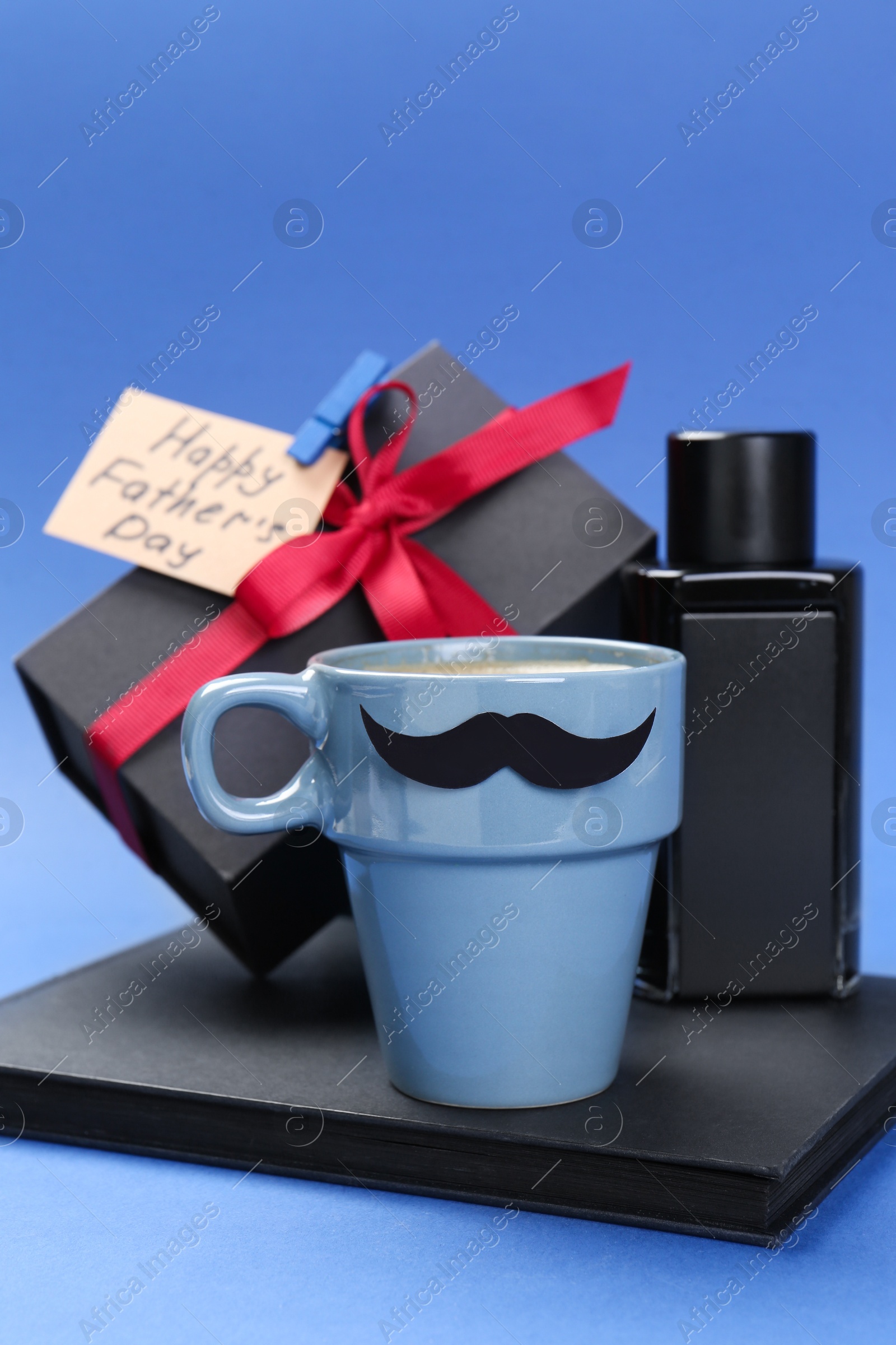 Photo of Happy Father's Day. Cup with paper mustache, notebook, perfume, card and gift box on blue background, closeup