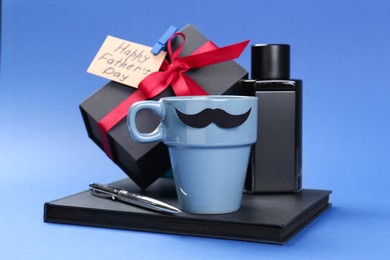 Photo of Happy Father's Day. Cup with paper mustache, notebook, pen, perfume, card and gift box on blue background, closeup