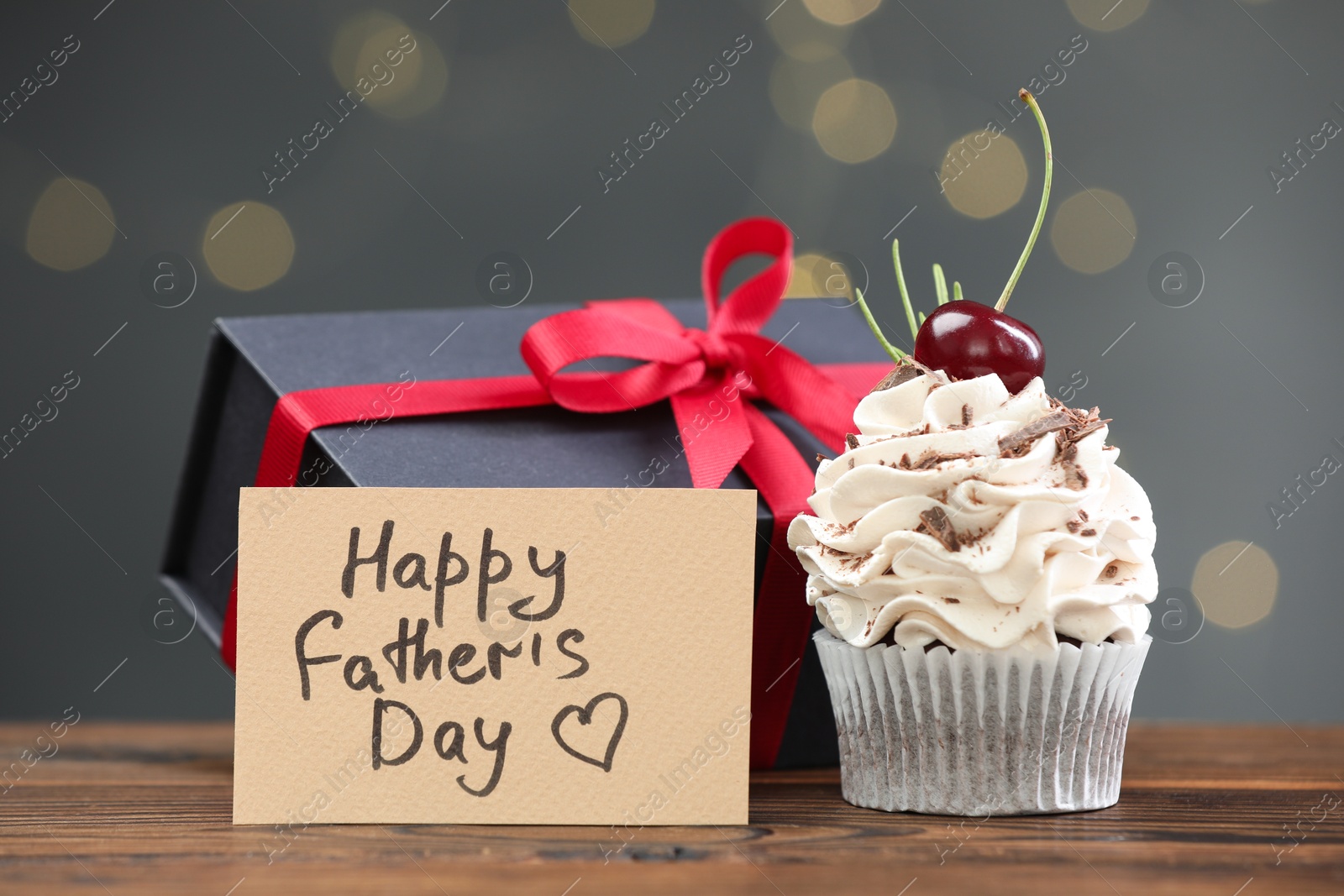 Photo of Greeting card with phrase Happy Father's Day, cupcake and gift box on wooden table against blurred lights