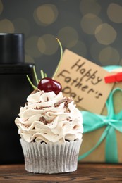 Photo of Greeting card with phrase Happy Father's Day, cupcake, gift box and perfume on wooden table against blurred lights, closeup