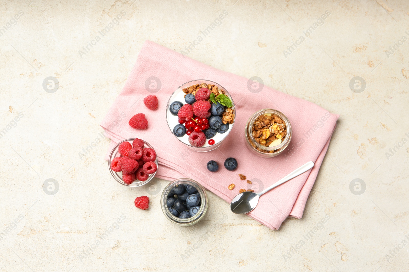 Photo of Tasty yogurt with fresh berries, granola, mint in glass and spoon on gray textured table, flat lay