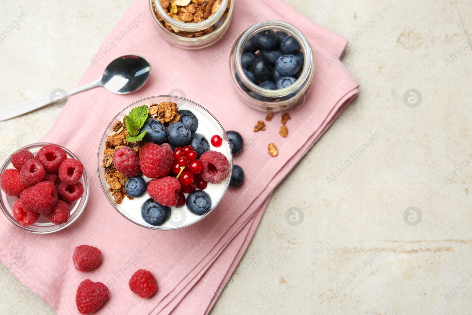 Photo of Tasty yogurt with fresh berries, granola, mint in glass and spoon on gray textured table, flat lay. Space for text