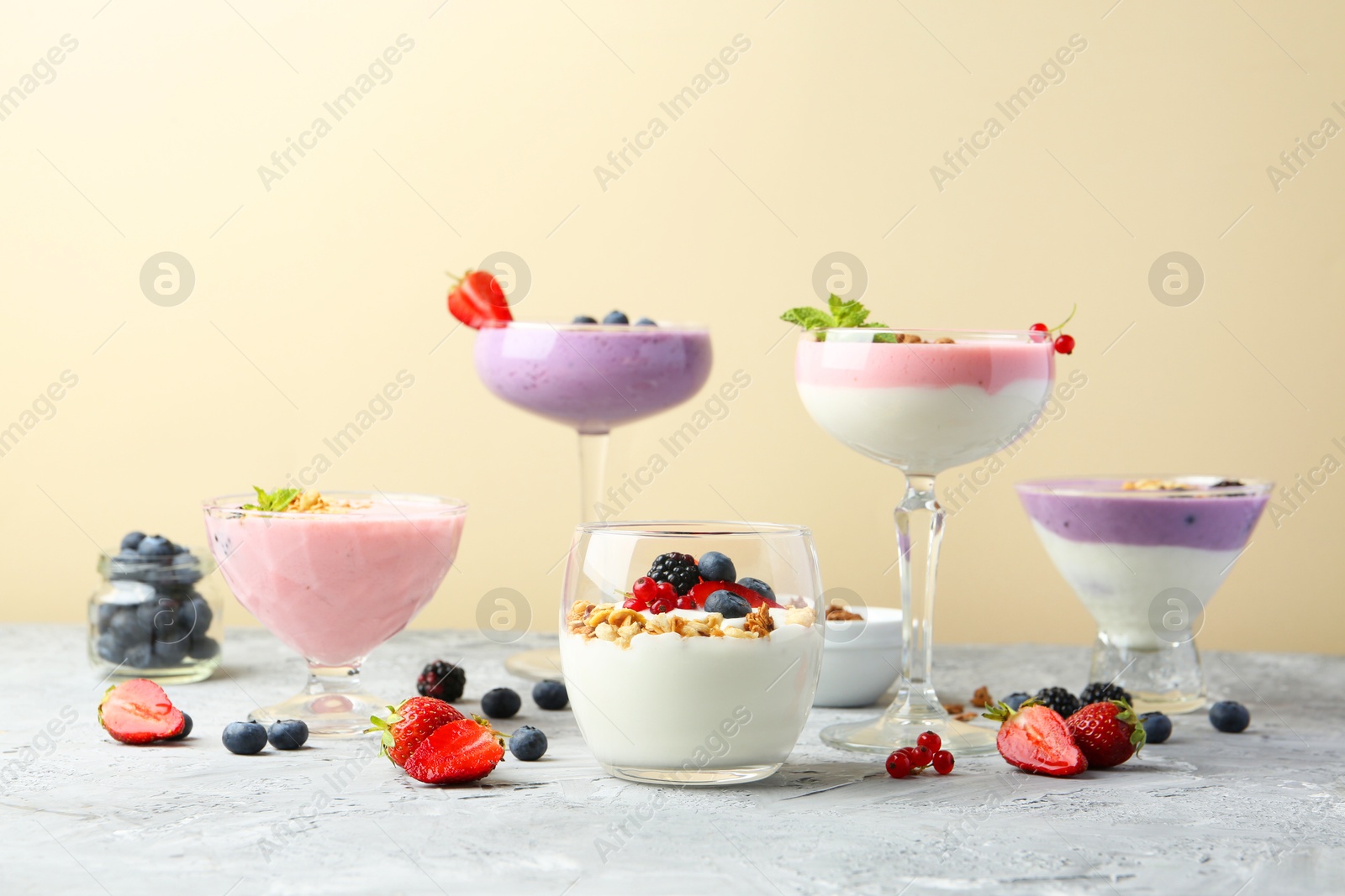 Photo of Different tasty yogurts with fresh berries and granola in glass dishware on gray textured table against pale yellow background