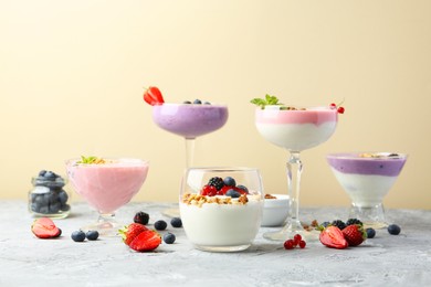 Different tasty yogurts with fresh berries and granola in glass dishware on gray textured table against pale yellow background