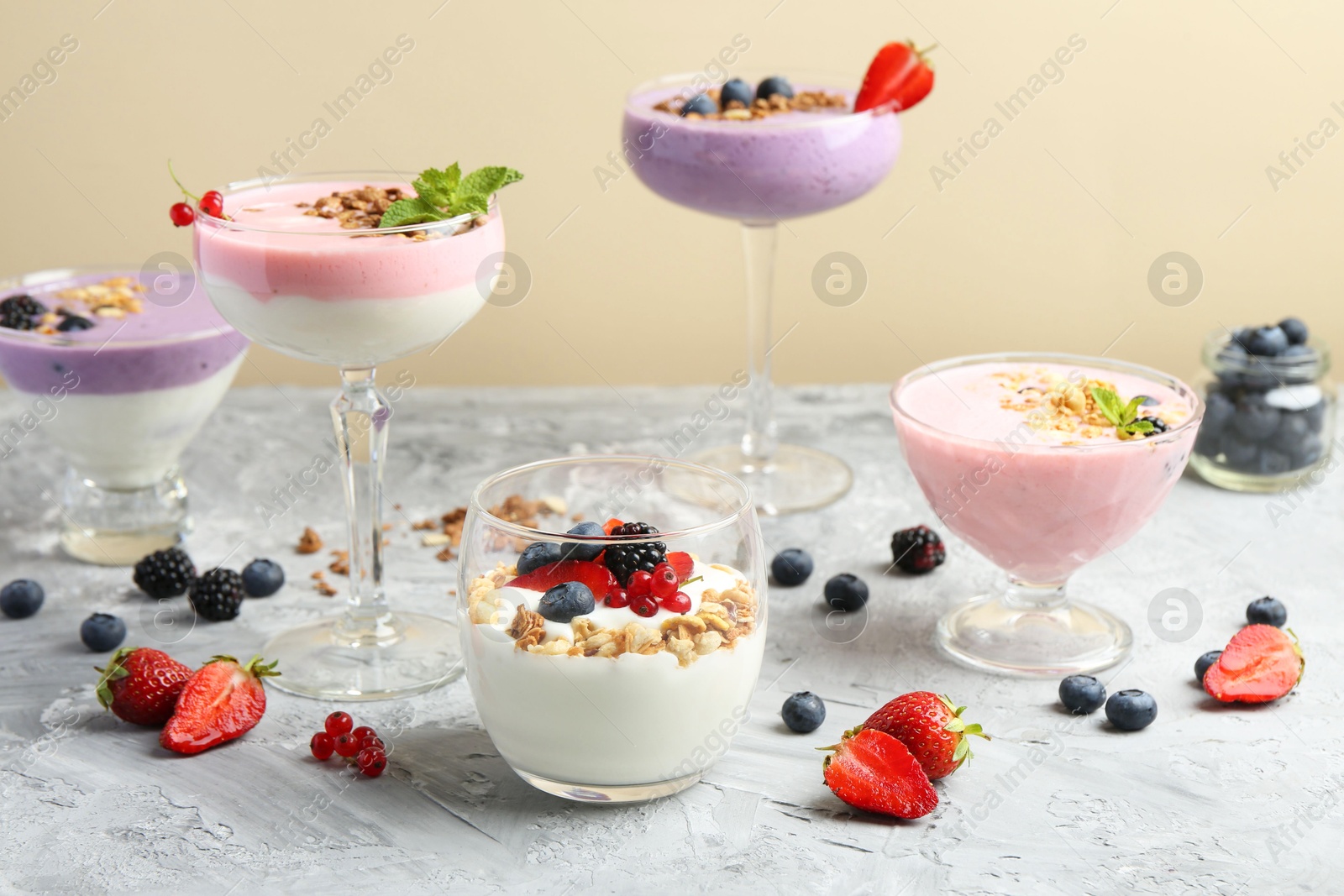 Photo of Different tasty yogurts with fresh berries and granola in glass dishware on gray textured table against pale yellow background