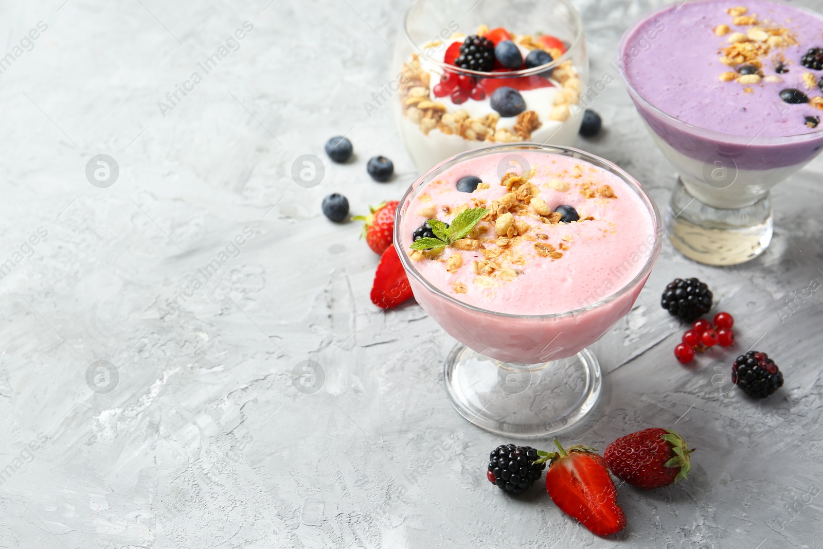 Photo of Different tasty yogurts with fresh berries and granola in glass dessert bowls on gray textured table, closeup. Space for text