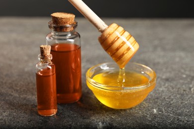 Photo of Pouring honey from dipper into bowl and bottles of tincture on grey textured table, closeup. Alternative medicine