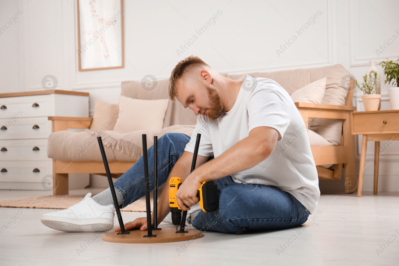 Photo of Man with electric screwdriver assembling table on floor in room