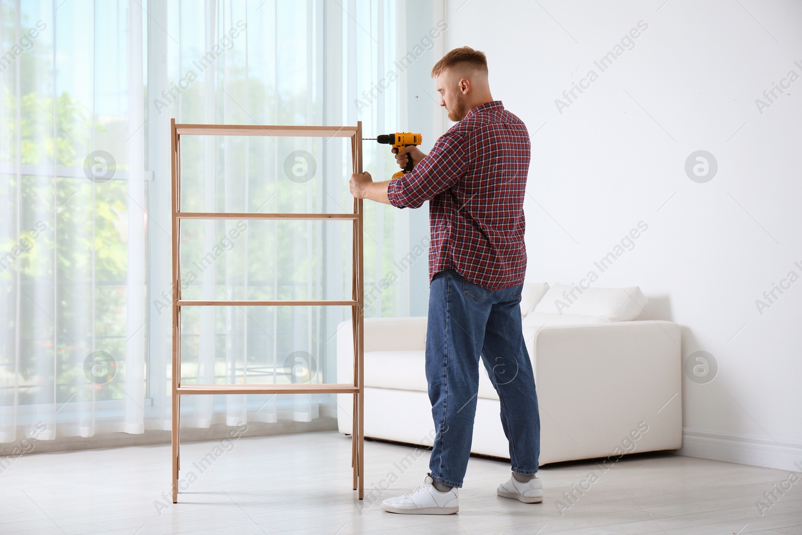 Photo of Man with electric screwdriver assembling wooden shelving unit in room