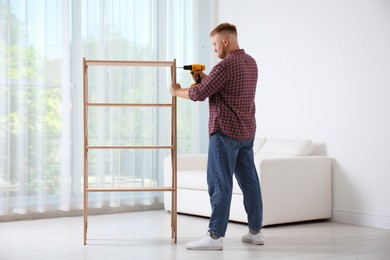 Photo of Man with electric screwdriver assembling wooden shelving unit in room