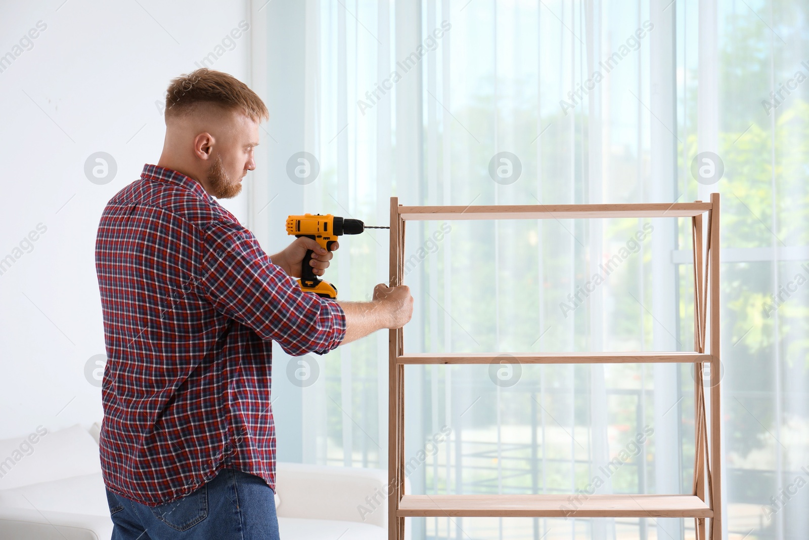 Photo of Man with electric screwdriver assembling wooden shelving unit in room