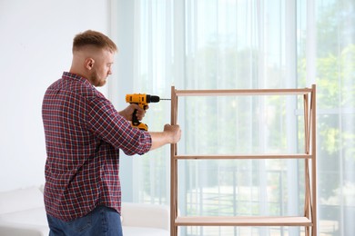 Man with electric screwdriver assembling wooden shelving unit in room