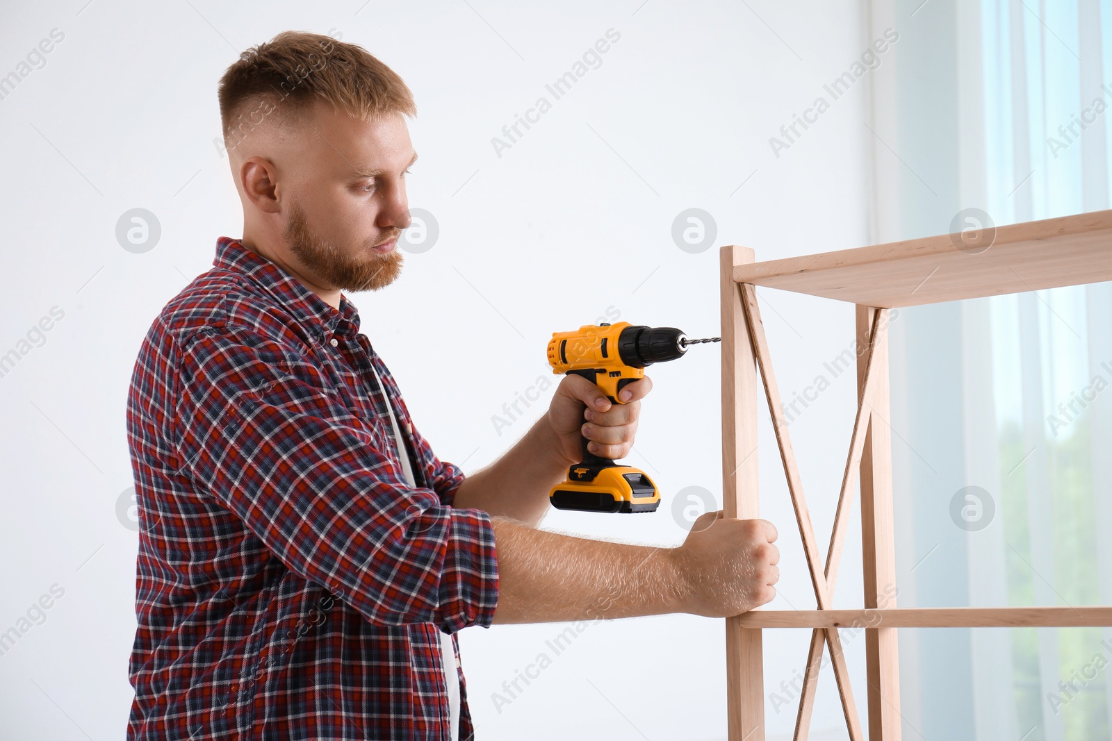 Photo of Man with electric screwdriver assembling wooden shelving unit in room