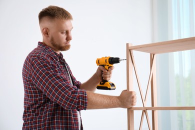 Photo of Man with electric screwdriver assembling wooden shelving unit in room