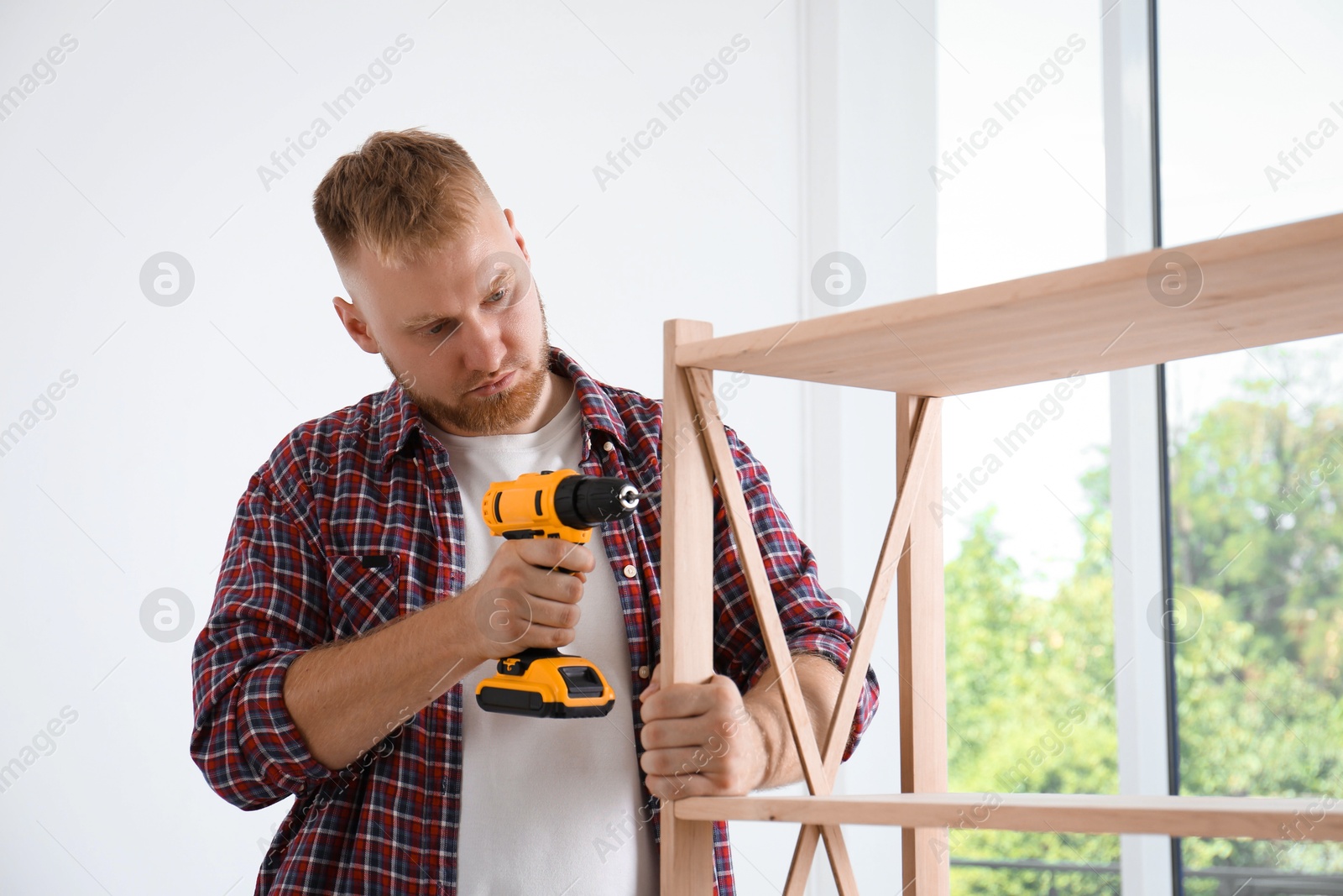 Photo of Man with electric screwdriver assembling wooden shelving unit at home