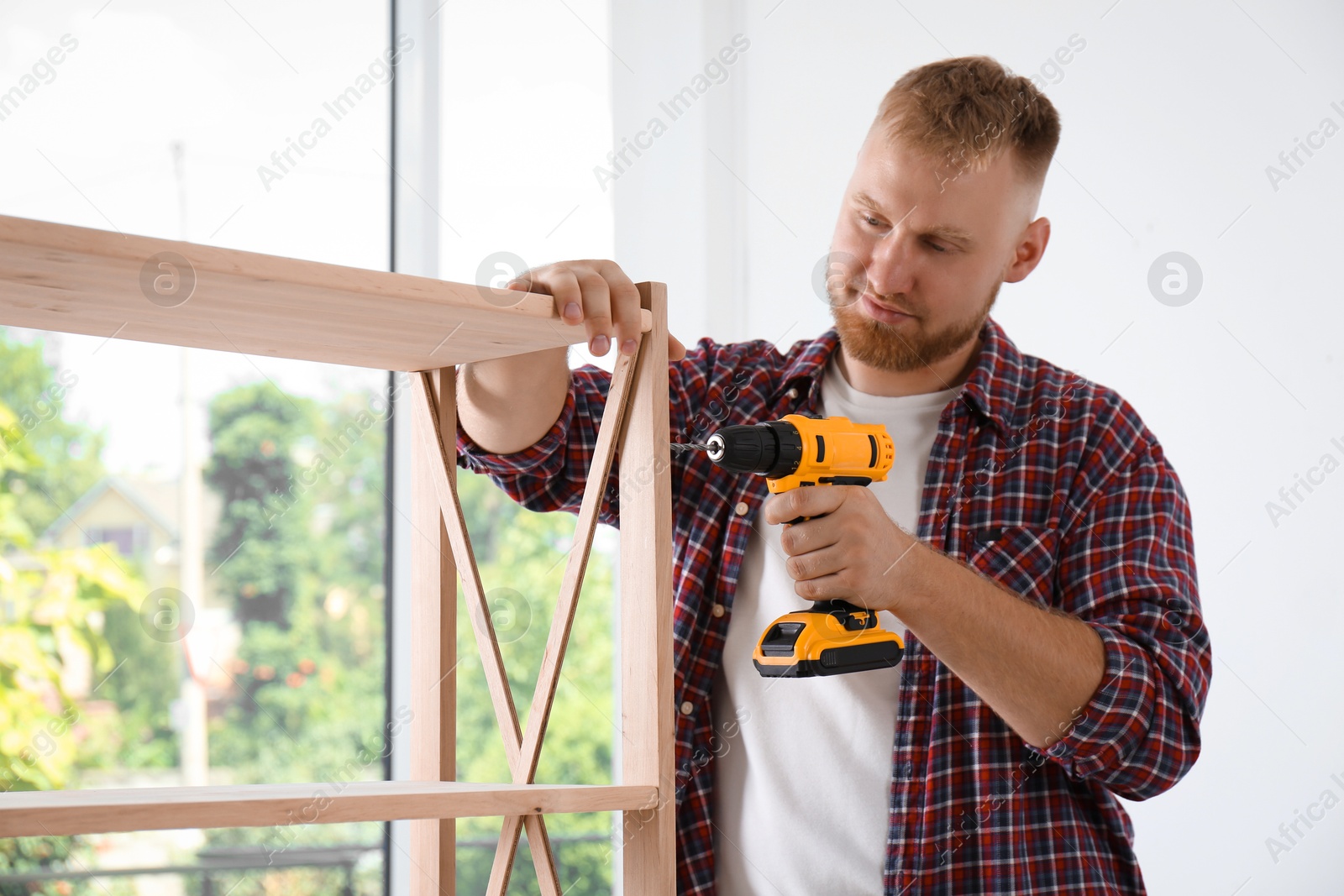 Photo of Man with electric screwdriver assembling wooden shelving unit at home